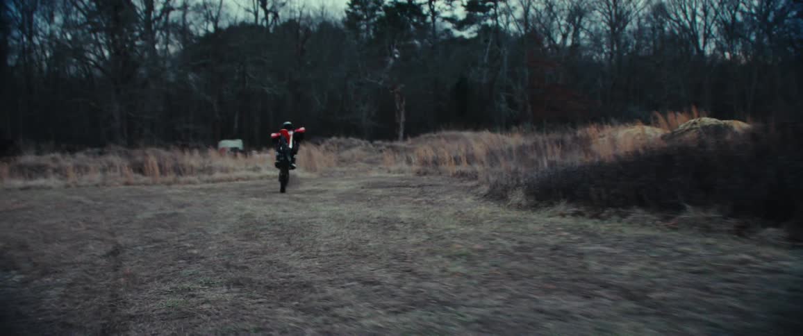 A person performs a jump on a red dirt bike over a red ramp in a grassy wooded area and then does a wheelie