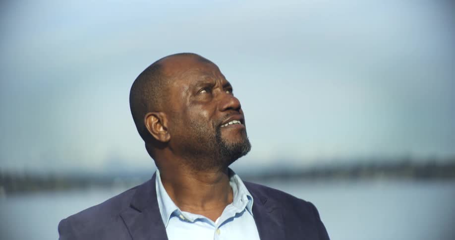 A black man in a blazer looks upwards facing skyward in an outdoor setting with soft sunlight and a blurred backdrop