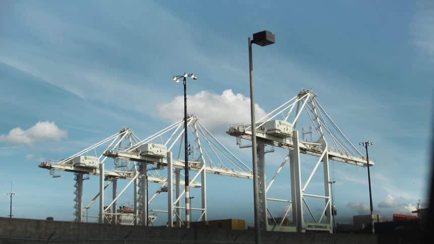 Three large white gantry cranes stand against a bright blue sky with scattered clouds in a port setting
