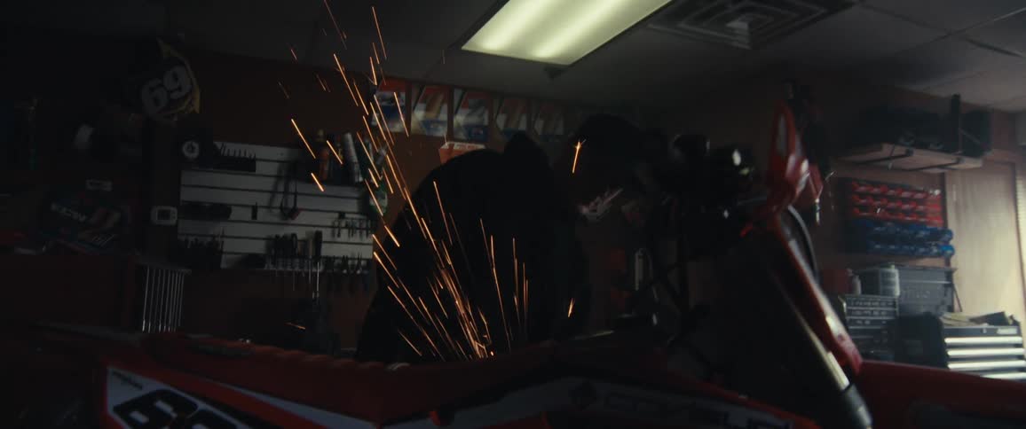 A person is using a power tool to grind or cut metal on a motorcycle causing bright sparks to fly in a workshop filled with various tools and equipment