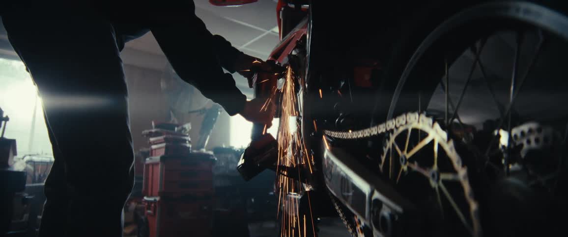 A person is using a power tool to grind or cut metal on a motorcycle causing bright sparks to fly in a workshop filled with various tools and equipment