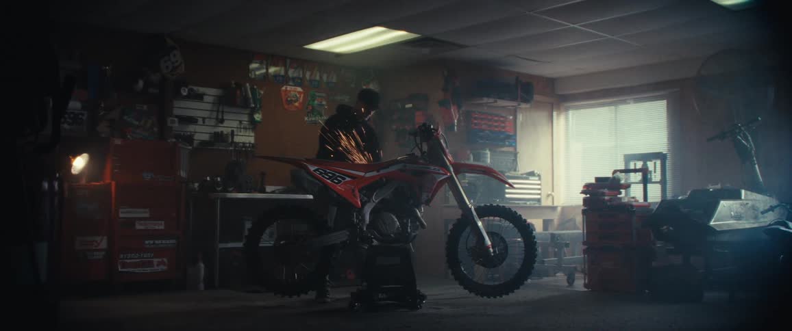 A person is working on a red and white dirt bike in a dimly lit garage with sparks flying from the area being worked on surrounded by various tools and equipment