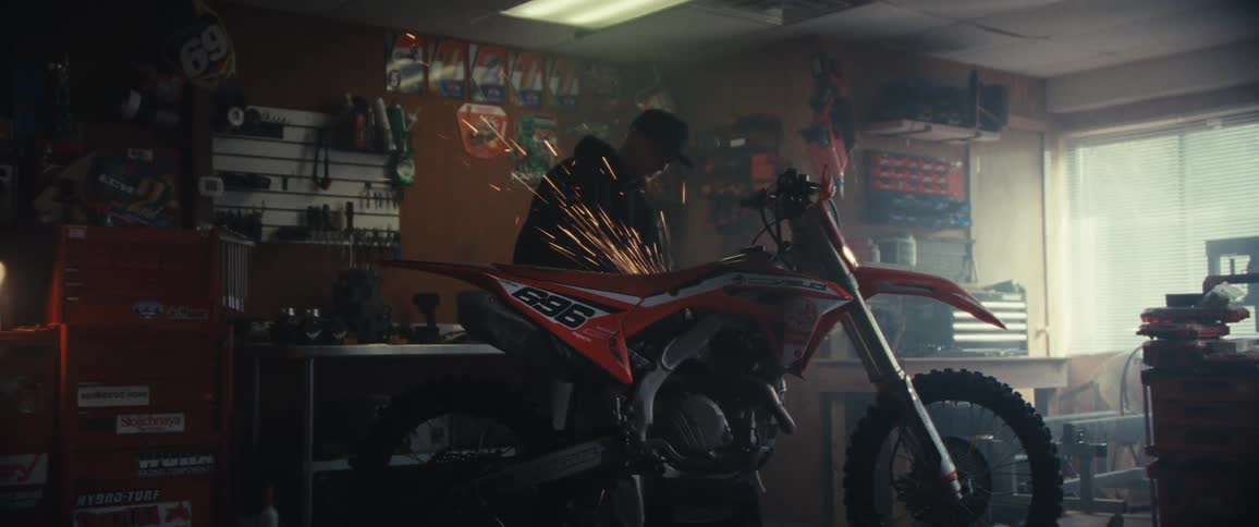 In a motorcycle shop a man grinds an orange bike as bright sparks fly off the side