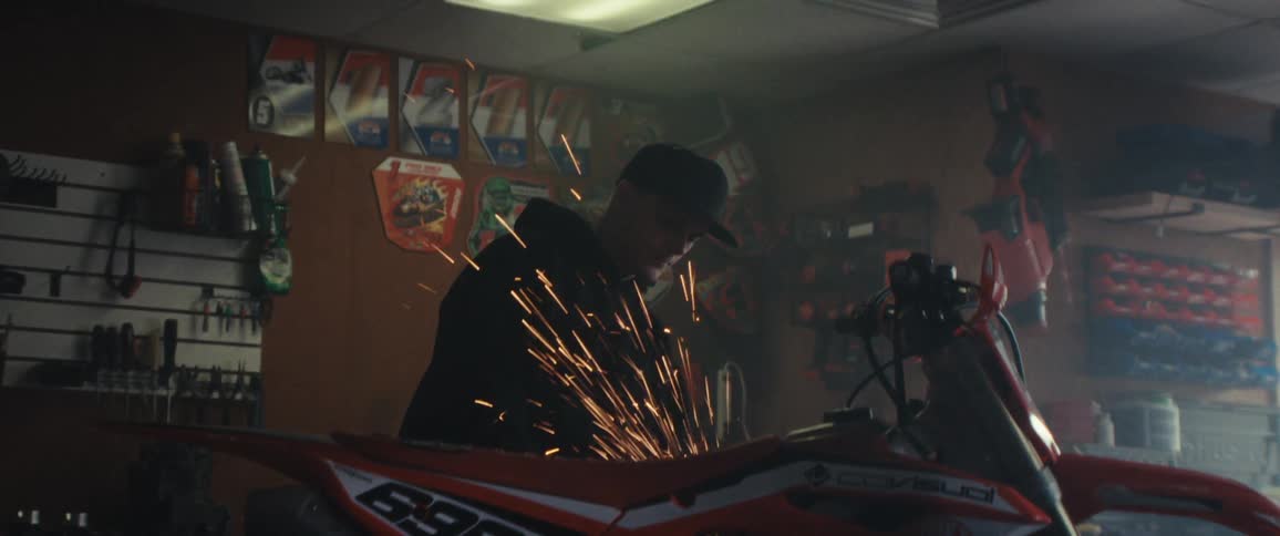 A person is using a power tool to grind or cut metal on a motorcycle causing bright sparks to fly in a workshop filled with various tools and equipment