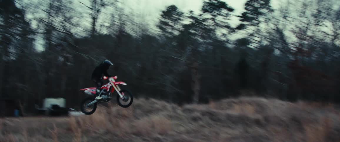 A person dangerously rides a motorcycle up a ramp placed on a grassy overgrown field
