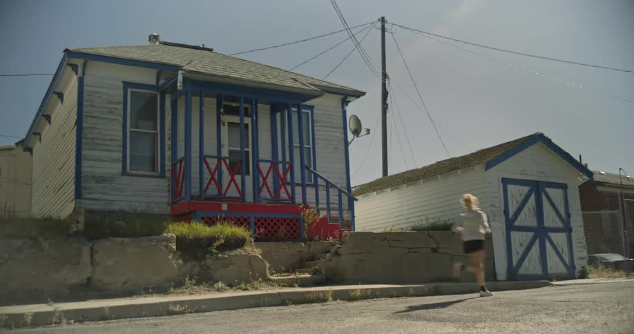 A woman runs along a sidewalk past a small weathered house with blue trim and a detached garage under a bright sky