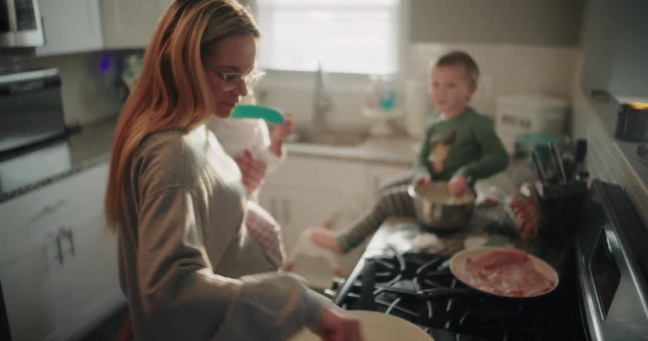 Mom holds daughter and playfully interacts with son who tastes pancake batter he mixes while mom stirs a pot near cooking meat