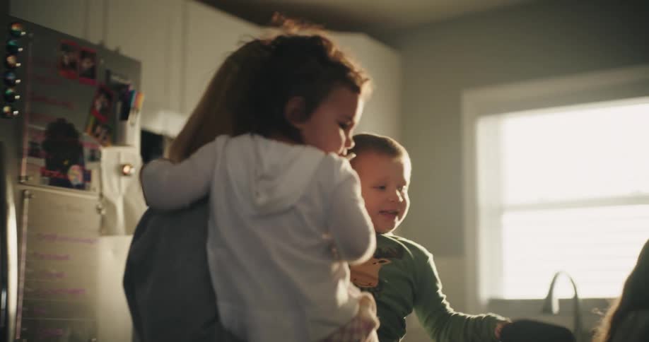 Mother in her kitchen lifts her younger children while an older daughter works at the counter