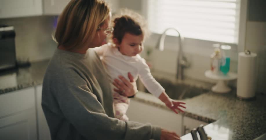 In a kitchen a mother holding her toddler daughter first offers an apple then a pacifier as the toddler reaches for a pastry on the counter