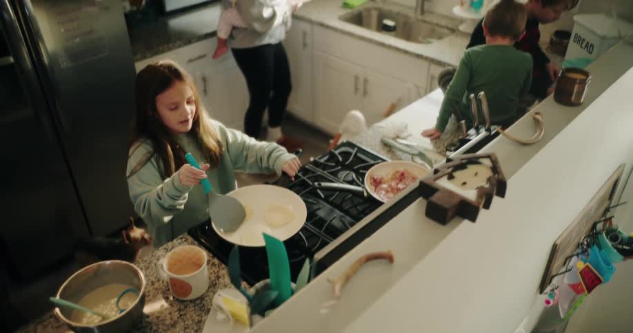 A young girl cooks pancakes on a stove in a busy kitchen scene with other children and an adult