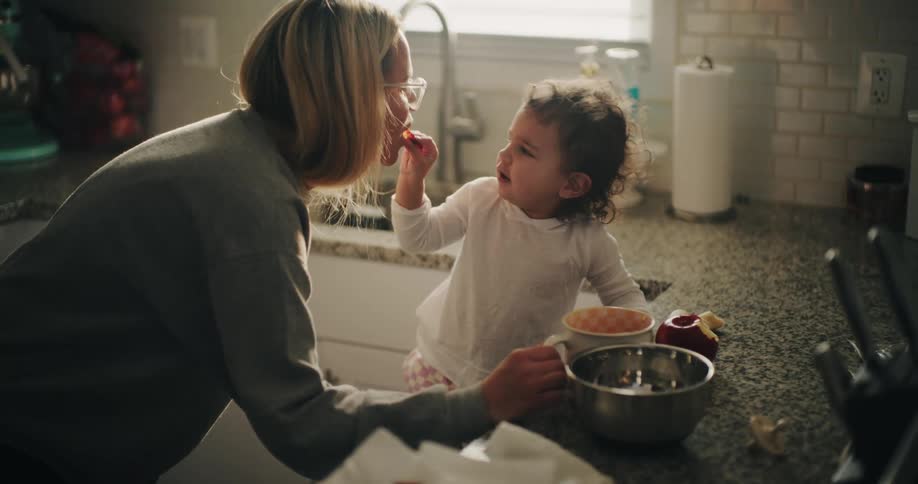 Mother kisses her daughter after the child shares food with her while they are in their home kitchen