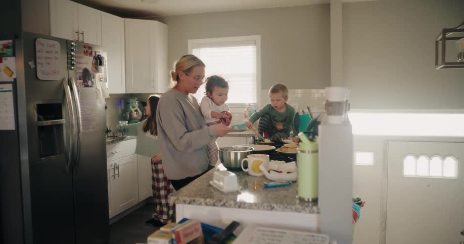 In a bright kitchen a mother cooks pancakes while holding her toddler daughter as her son mixes batter and her older daughter closes the fridge showcasing family preparing breakfast