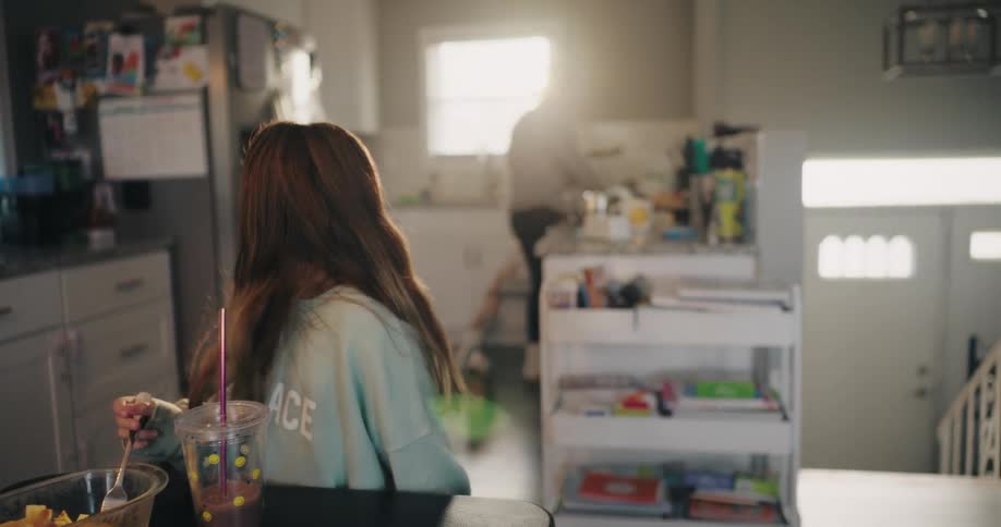 In a bustling kitchen a mother cooks while holding a toddler as her son assists and her daughter eats breakfast at the foreground table bathed in sunlight