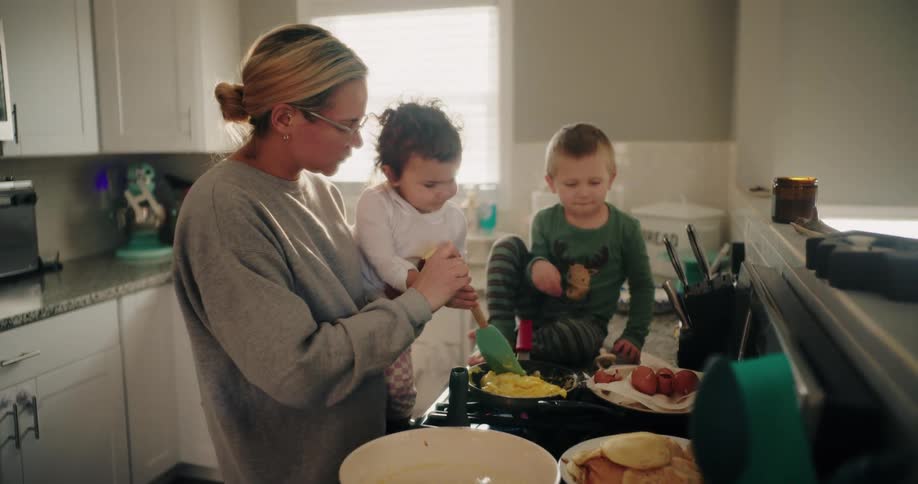 A mother holds a child and serves food on a plate in a kitchen while another child stands by the sink with plates of food on the counter