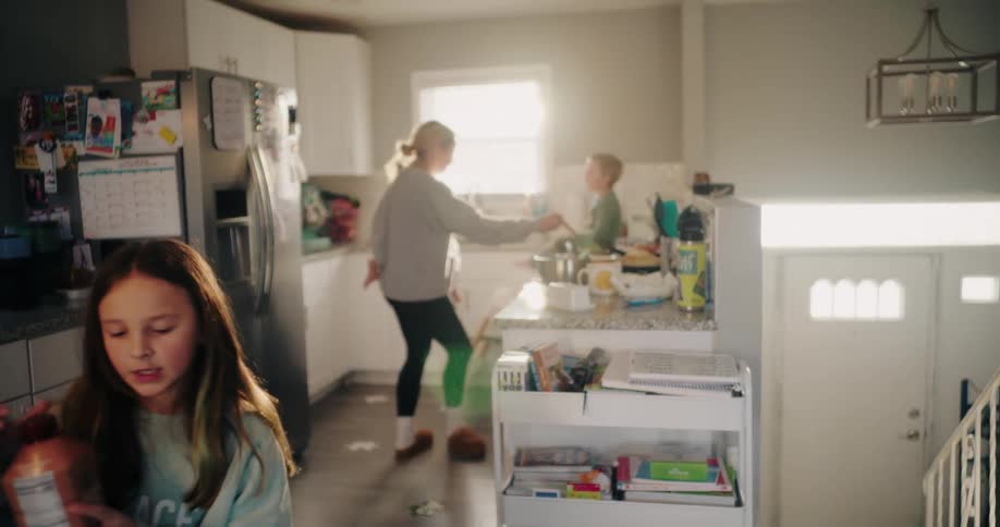 A mother prepares food in the kitchen while holding a child another child stands by the sink and her daughter eats at the table
