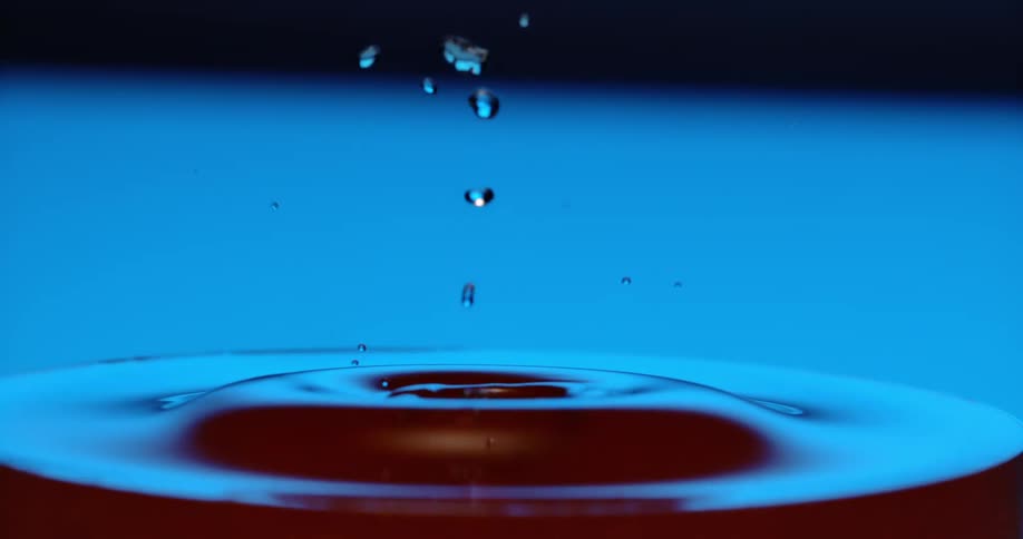 A droplet of falls into a glass of water creating a splash and concentric ripples on the surface all set against a blue background