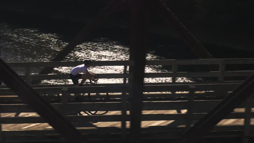 A cyclist in a white shirt rides across a wooden bridge on a sunny day