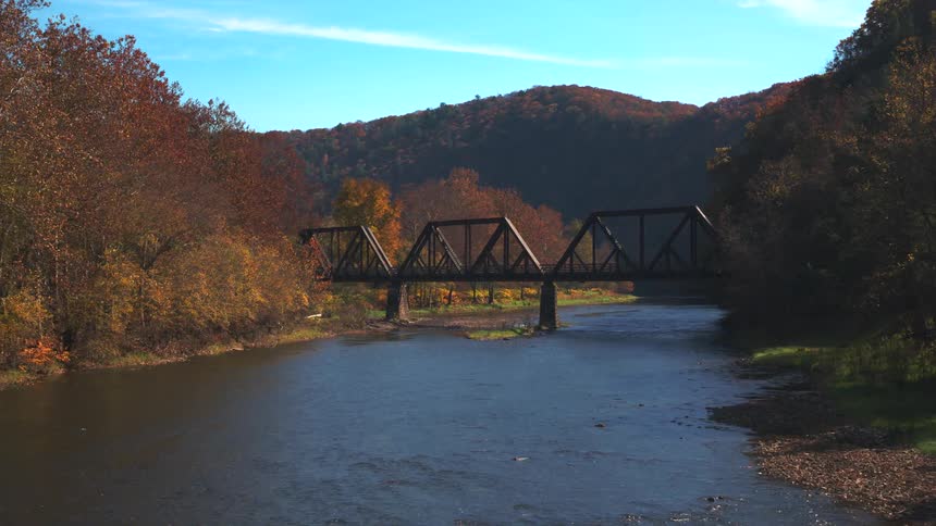 Flying above a river towards an old bridge with autumncolored trees lining the riverbanks and distant orange hills