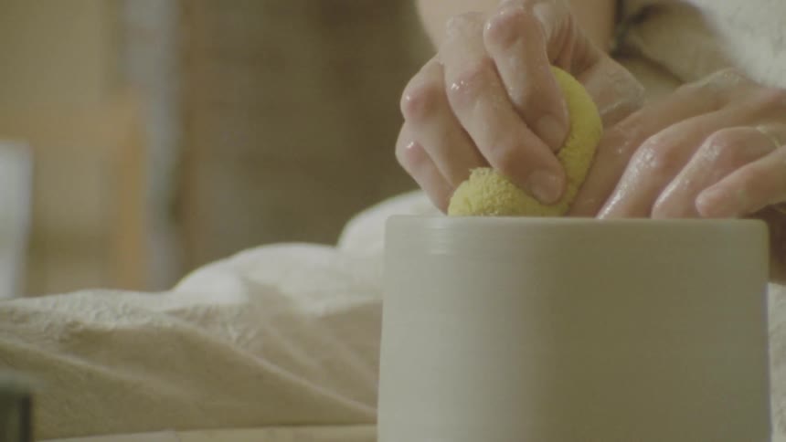 A persons hands press a yellow sponge into a white ceramic container on a pottery wheel