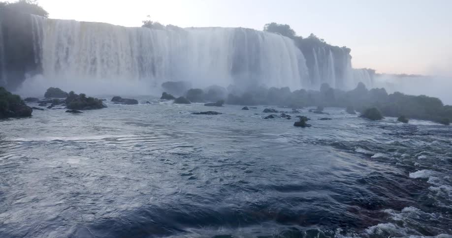 A wide multitiered waterfall cascades over a rocky cliff surrounded by lush forest with mist above the falls