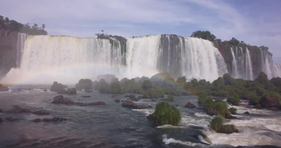 A wide multitiered waterfall cascades over a rocky cliff surrounded by lush forest with mist above the falls