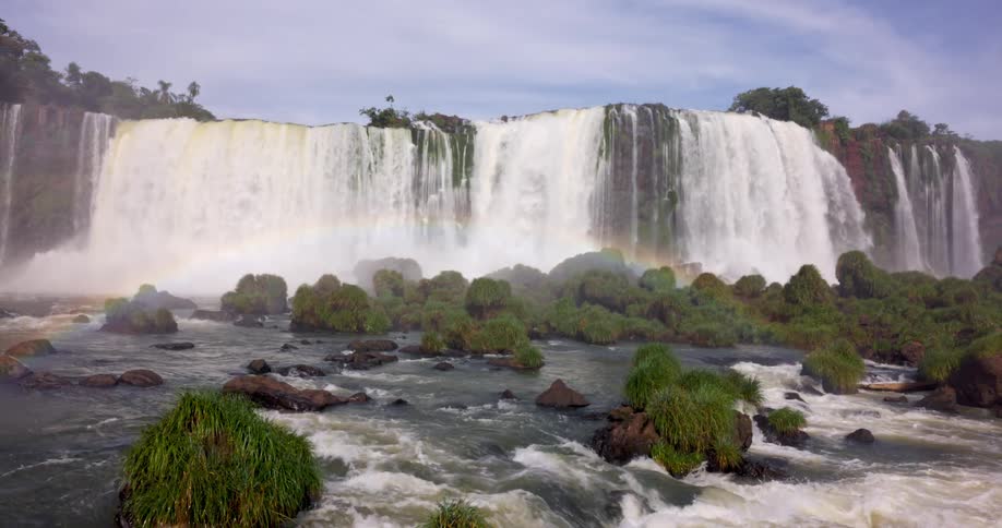 A wide multitiered waterfall cascades over a rocky cliff surrounded by lush forest with a faint rainbow visible in the mist above the falls