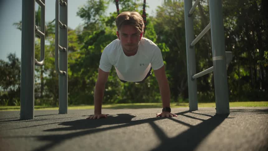 A man is doing pushups outdoors with his feet in gymnastics hoops in a fitness area