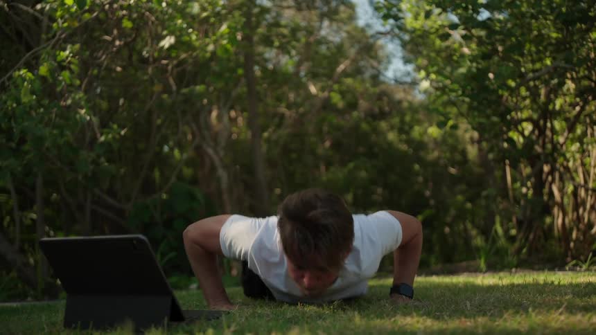 A man is crouched down looking at a laptop screen and then begins doing pushups on a grassy floor outside
