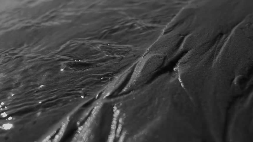Water flows over dark textured sand creating intricate patterns and reflections