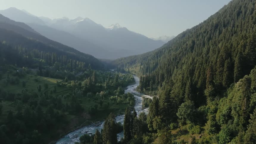 A winding river flows through a lush forested valley flanked by steep treecovered mountains with distant snowcapped peaks visible under a clear sky