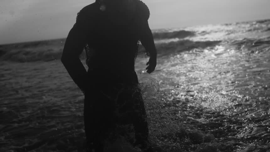In slow motion a silhouetted man in a netlike garment runs through ocean waves water splashing around him