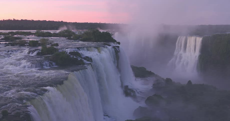 A wide multitiered waterfall cascades over a rocky cliff surrounded by lush forest with mist above the falls