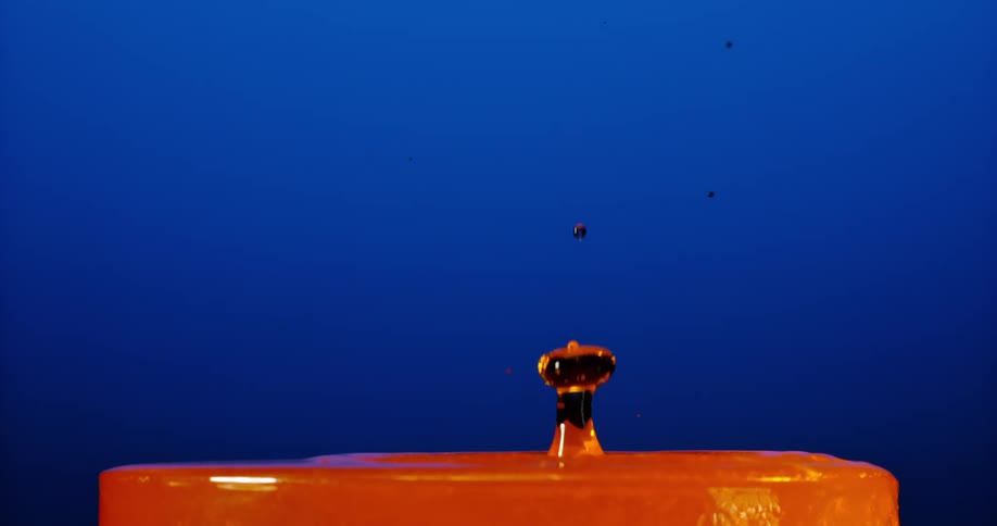 An orange liquid surface is disturbed by a falling drop against a blue background resulting in a splash and a stem like formation
