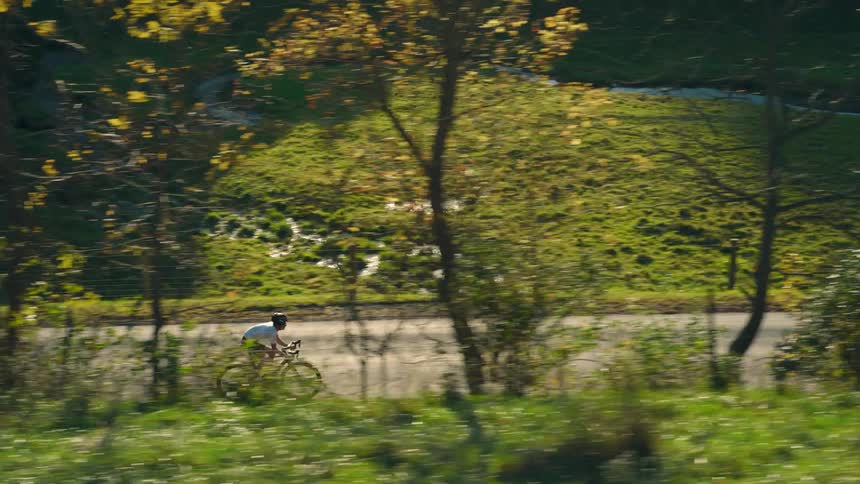 A person in a red outfit and yellow hat a cyclist wearing a helmet and white shirt is riding a bicycle on a road surrounded by trees with autumn foliage and a grassy hill in the backgroundon a rocky shore facing a massive wave crashing against the rocks