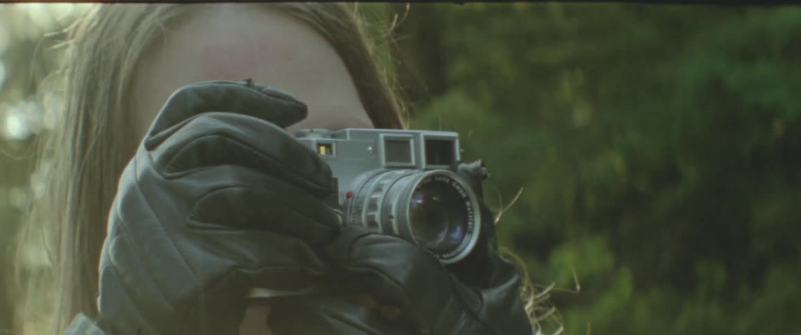 A person with long hair and wearing leather gloves holds up a leica camera to take a picture
