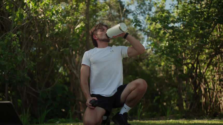 Man half kneels in a field and takes a drink from a container while recovering from exercise