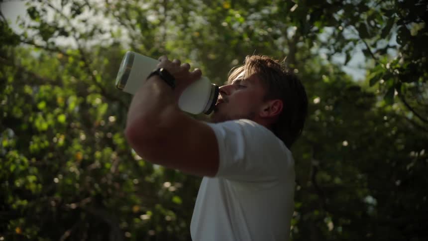 A man in a white t shirt takes a drink from a metal water bottle