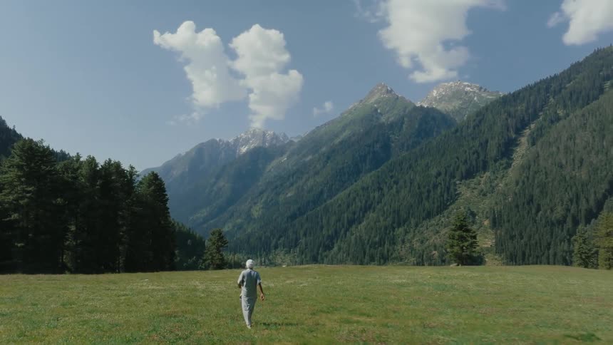 A man in a white turban and long indian shirt walks across a green grass field toward a massive treecovered mountain