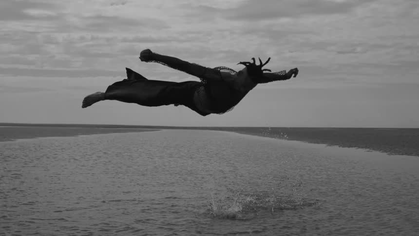 Man wearing a wide legged outfit floats in mid air over the water on an overcast day