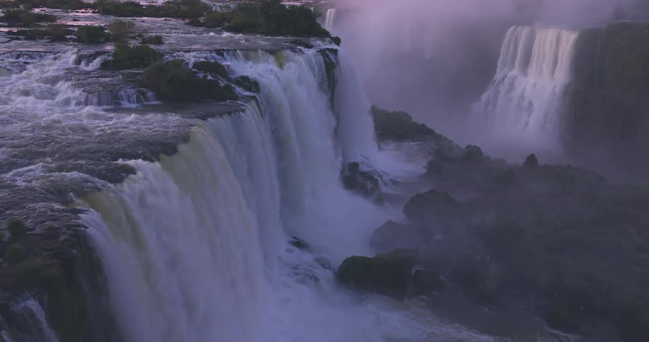 A wide multitiered waterfall cascades over a rocky cliff surrounded by lush forest with mist above the falls