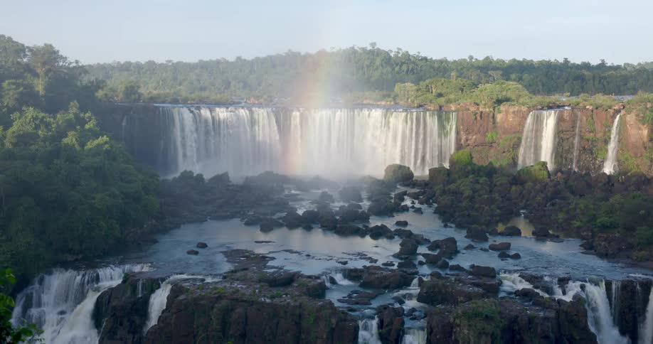 A wide multitiered waterfall cascades over a rocky cliff surrounded by lush forest with a faint rainbow visible in the mist above the falls
