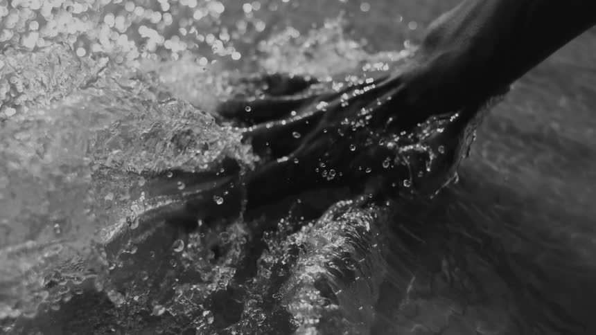 A persons hand disrupts the waters flow swiping upwards and creating a splash of water droplets in black and white
