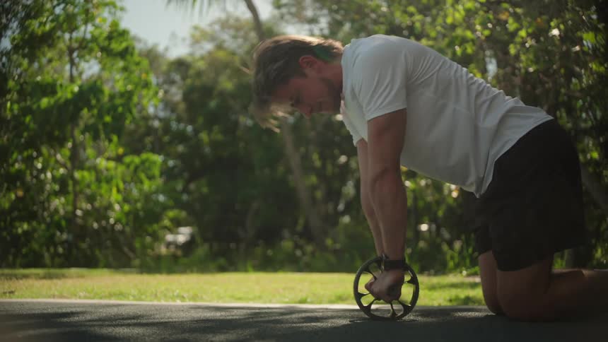 A man is performing an ab wheel exercise on an outdoor path extending their body forward with arms fully stretched while kneeling on the ground surrounded by lush green trees and sunlight