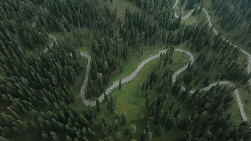 An aerial view reveals a winding mountain pass through a dense forest with cars navigating the scenic road as the perspective rises