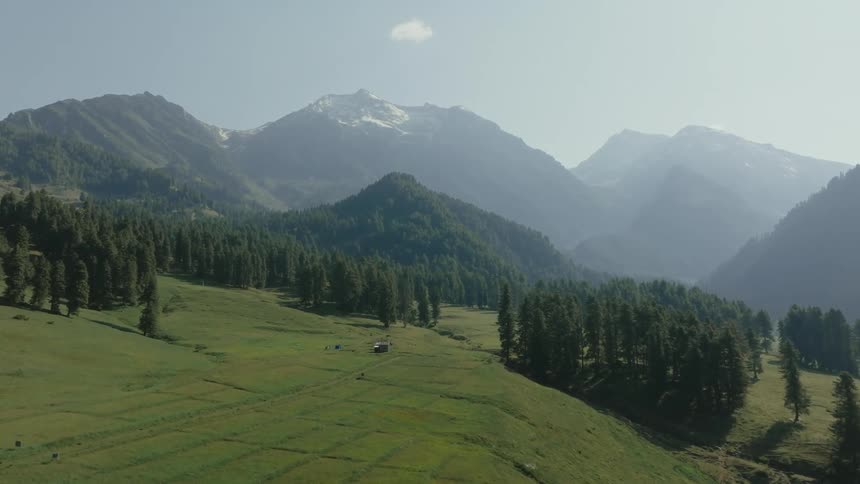Aerial view of a hillside with a small shed and grazing livestock set against a mountain backdrop