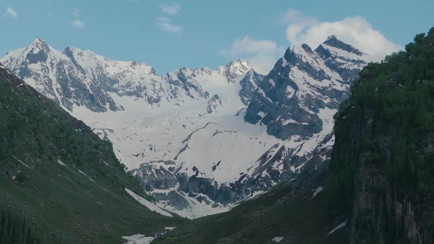 A mountain landscape with snowcovered peaks and rugged cliffs flanked by green valleys and patches of snow under a clear blue sky