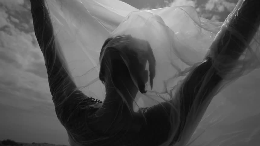 A person is standing with his back turned with a transparent sheet around his body and with his arms raised up and everything is in black and white tones