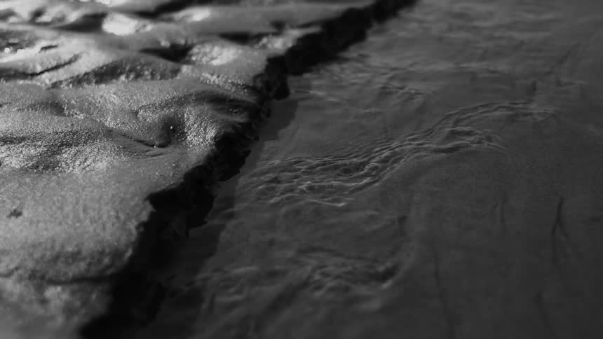 Black and white close up shows the texture of sand meeting rippling water