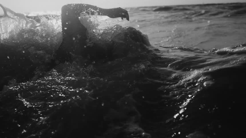 A person is swimming in the ocean with an arm raised above the water creating splashes and ripples in the sunlight