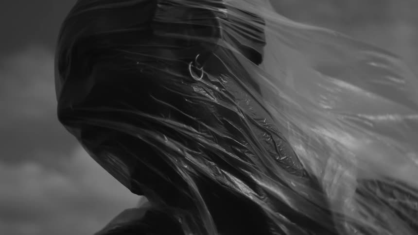 The face of a person wrapped in plastic with the plastic billowing in the wind set against a cloudy sky